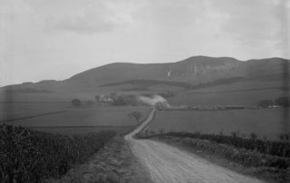 Image shows an old photograph taken during the 1970's of Swanston Farm with the Pentland Hills in the background. There is a smudge of smoke rising from one of the farm cottages in the centre.