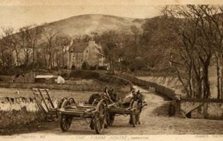Image shows a very old postcard of a black and white photograph taken in the 1800's by James Patrick of the Farm House at Swanston. The white farmhouse is partially hidden in the trees in the background, with two carthorses hitched-up to carts in the foreground, being led by a farm hand up the track towards the house. Some parts from the horse's carts are leaning up against the stone wall on the left in the foreground.