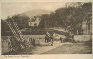 Image shows a very old postcard of a black and white photograph taken in the 1800's by James Patrick of the Farm House at Swanston. The white farmhouse is partially hidden in the trees in the background, with two Clydesdale horses in the foreground, one being ridden and the rider leading the second one, walking up the track towards the house. The horse's carts are leaning up against the stone wall on the left in the foreground, and there is a gentleman in a suit standing with his hands in his pockets beyond the horses, as though he is waiting for them.
