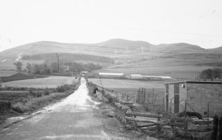 Image shows an old photo taken in 1975 of Swanston Farm, before the City Bypass was built (which was in 1981). The old wooden clubhouse for the golf club and the farm buildings are visible as well as the T-Wood with Caerketton Hill in the background.