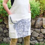 Photograph shows a full-length cream linen apron (picture cuts it off below the chest) worn by a man. The head of a highland cow is printed on the bottom of the apron and there are 2 deep pockets to the front, level with the thighs of the model. The model has his hands in these pockets. There is a green hedge and a stone wall behind the model.