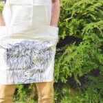 Photograph shows a full-length cream linen apron (picture cuts it off below the chest) worn by a man. The head of a highland cow is printed on the bottom of the apron and there are 2 deep pockets to the front, level with the thighs of the model. The model has his hands in these pockets. There is a green hedge behind the model.