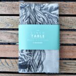 Photograph shows a folded pack of 4 cream linen napkins wrapped in a teal-coloured card label saying 'The Linen Table. Woven in Scotland. 4 Napkins' on it. The napkins all have highland cow heads printed on them. They are displayed on a wooden table.