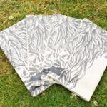 Photograph shows 4 folded cream linen napkins with highland cow heads printed on them. They are displayed in a fan-shape on a grass background.