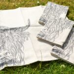 Photograph shows 3 folded and one open cream linen napkins with highland cow heads printed on them. They are displayed on a grass background.
