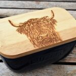 Photograph shows a Navy blue ceramic butter dish with a wooden lid. The dish has the word 'Butter' embossed on the side. The lid has a highland cow head etched on the top. The butter dish is displayed on a wooden table.