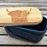 Photograph shows a Navy blue ceramic butter dish with a wooden lid, slightly off-set for display purposes so you can see inside the item. The butter dish has the word 'Butter' embossed on the side. The lid has a highland cow head etched on the top. The butter dish is displayed on a wooden table.