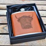 Photograph shows a brown leather hip flash with metal lid and the head of a Highland Cow etched on the leather on the front. It is shown displayed in its black presentation box on a wooden table.