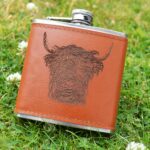vPhotograph shows a brown leather hip flash with metal lid and the head of a Highland Cow etched on the leather on the front. It is shown displayed on a grass background