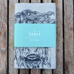Photograph is a cream-coloured linen table-runner, folded with a teal-coloured card label saying 'The Linen Table. Woven in Scotland. Table Runner' on it and with a highland cow head printed on it the runner. The table runner is lying on a wooden table for display.