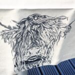 Photograph is a cream-coloured linen table-runner with a highland cow head printed on it. The table runner is lying on a table for display.