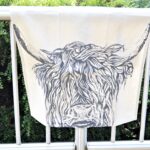 Photograph is a cream-coloured linen tea towel with a highland cow head printed on it. The tea towel is draped over a metal railing with a green hedge behind it for display.