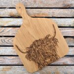 Photograph is of a wooden chopping board with curved corners and with a wooden handle and with a highland cow head etched on it. The board is lying on a wooden table.