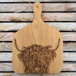 Photograph is a close-up of a wooden chopping board with curved corners and with a wooden handle and with a highland cow head etched on it. The board is lying on a wooden table.