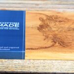 Photograph is a close-up of a wooden chopping board with a wooden handle and with a highland cow head etched on it, with a blue card label saying 'Scottish Made'. The board is lying on a wooden table.