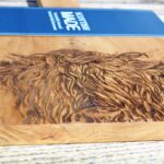 Photograph is a close-up of a wooden chopping board with a wooden handle and with a highland cow head etched on it, with a blue card label saying 'Scottish Made'. The board is lying on a wooden table.