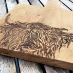 Photograph is a close-up of a wooden chopping board with a wooden handle and with a highland cow head etched on it. The board is lying on a wooden table.