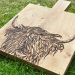 Photograph is of a wooden chopping board with a wooden handle and with a highland cow head etched on it. The board is lying on a grass background.