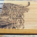 Photograph shows a close-up end of a wooden chopping board with a highland cow head etched on it. It is lying on a wooden table.