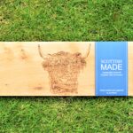 Photograph shows a long thin wooden tray with the head of a highland cow etched on it. It also shows both of the black metal handles at either end, and a blue card label wrapped round the board with 'Scottish Made' written on it. The board is on a grass background.
