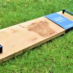 Photograph shows a long thin wooden tray with the head of a highland cow etched on it. It also shows both of the black metal handles at either end, and a blue card label wrapped round the board with 'Scottish Made' written on it. The board is on a grass background.