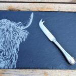 Photograph shows a slate cheeseboard with a Highland Cow head etched on it, with a silver cheese knife lying across it, displayed on a wooden table