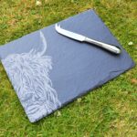 Photograph shows a slate cheeseboard with a Highland Cow head etched on it, with a silver cheese knife lying across it, displayed against a grass background