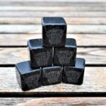 Photograph shows a pyramid stack of 6 slate cubed whisky stones with tiny Highland Cow heads etched on to them, on a wooden table