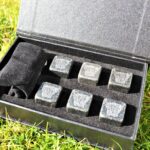Photograph shows a set of 6 slate cubed whisky stones with tiny Highland Cow heads etched on to them in a display box lying on grass