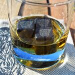 Image shows 3 slate cubed whisky stones with tiny Highland Cow heads etched on to them, in a glass of whisky, on a linen coaster on a wooden table