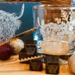 Image shows a variety of Highland Cow themed gifts including slate whisky stones, whisky glass, ice tongs, slate coaster and a wooden gift box in the background, all surrounded by Christmas decorations.